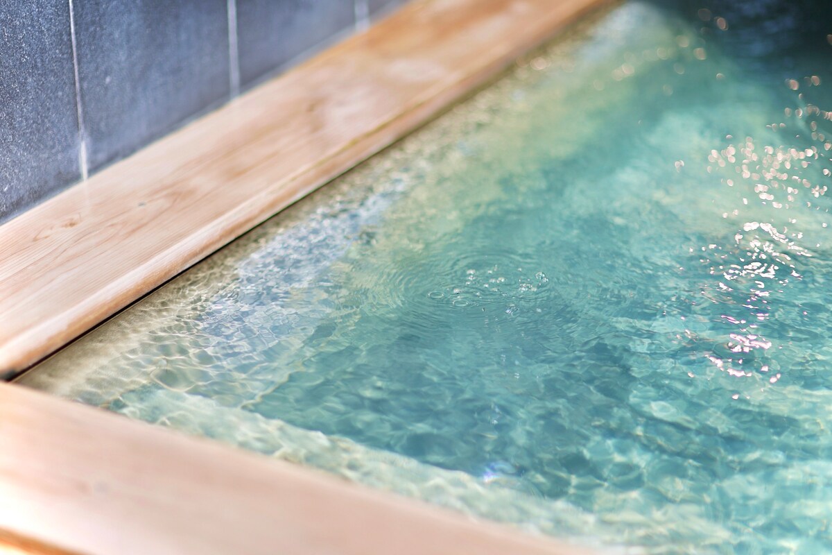 A tranquil onsen with clear blue water is framed by a smooth wooden edge. The gentle ripples in the surface reflect soft light, creating a peaceful ambiance. A simple stone wall serves as a backdrop, enhancing the serene atmosphere of the bathing area.