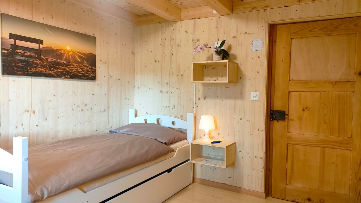 A cozy sleeping area features a simple, single bed with a beige comforter. The wooden walls and beams provide a natural feel, while a small shelf holds a lamp and a decorative item. A landscape photograph adorns one wall, enhancing the tranquility of the space.