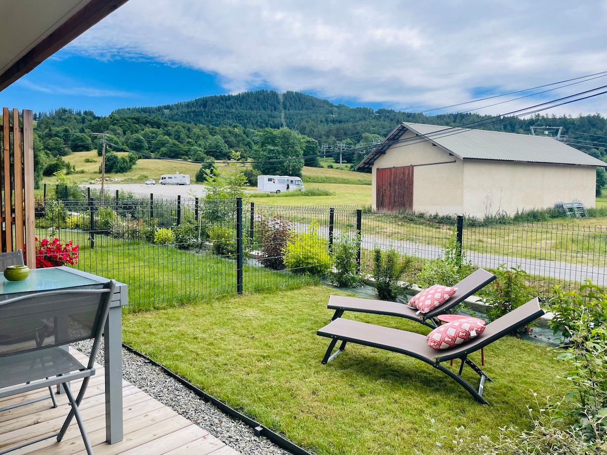 A private terrace is shown with two loungers and decorative cushions. Lush green grass is visible, bordered by a fence. Rolling hills and a distant outbuilding are seen in the background, creating a serene outdoor space ideal for relaxation.