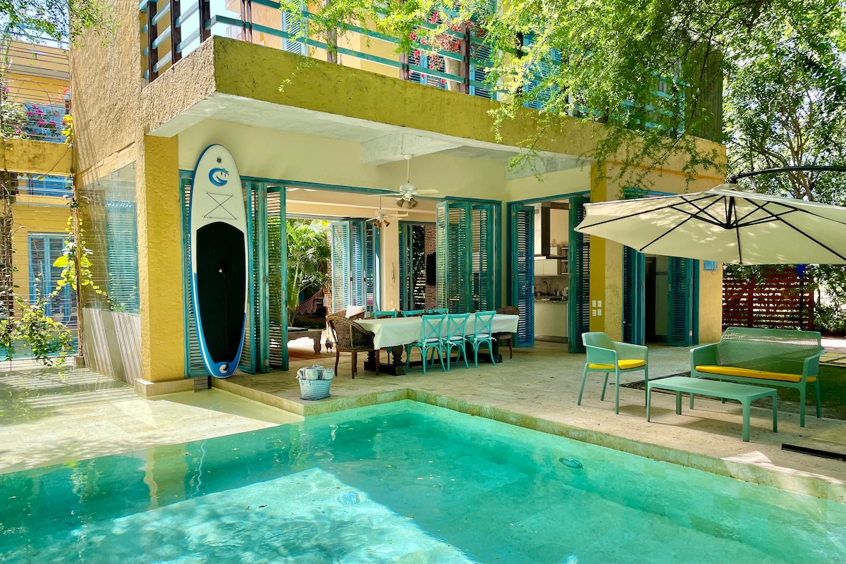 An outdoor area features a refreshing turquoise pool surrounded by natural foliage. A dining table with blue chairs is set under an umbrella, while a paddleboard is mounted on the wall beside the open, airy living space, enhancing the connection with the outdoors.
