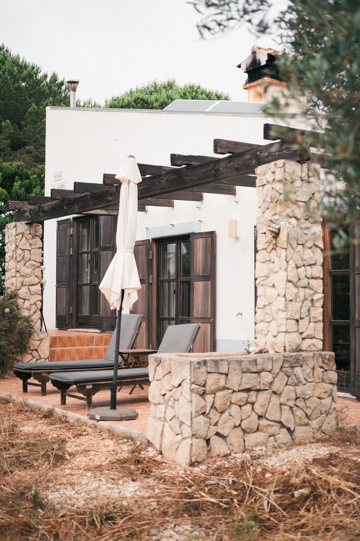An outdoor seating area is framed by a stone wall and wooden pergola, offering two lounge chairs positioned for relaxation. Sunlight filters through the leaves of nearby trees, and an umbrella provides shade, enhancing the calm ambiance of the space.