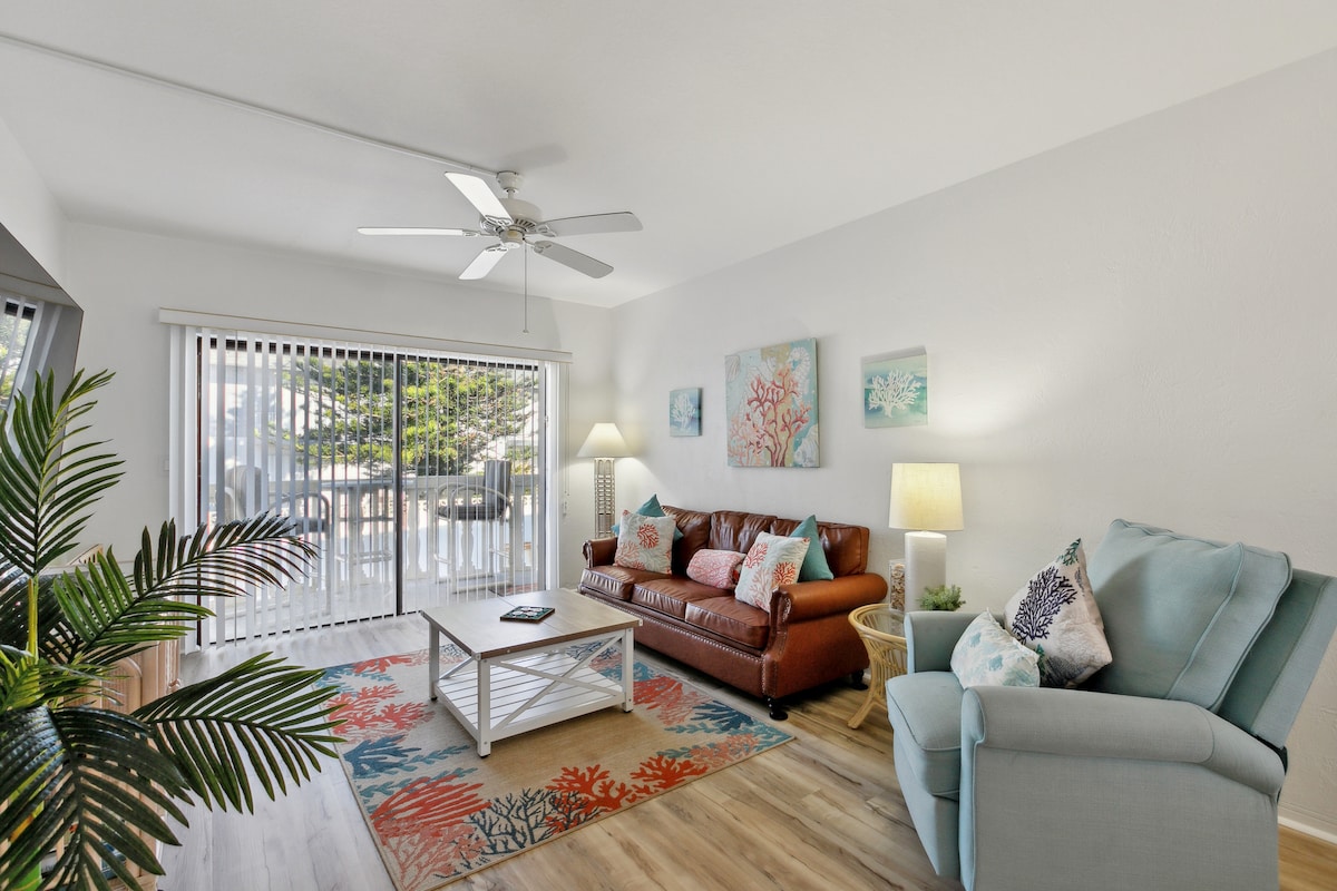 The living area is furnished with a brown leather sofa and a light blue armchair, accompanied by a coffee table on a colorful rug. Bright wall art and a ceiling fan complement the space, which features sliding glass doors that provide access to natural light.