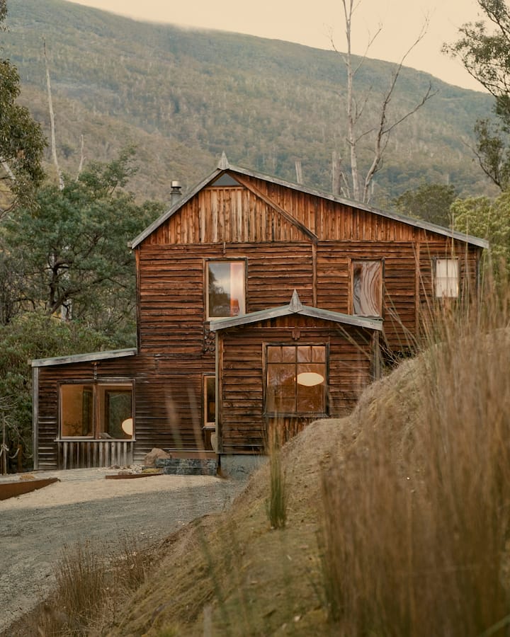 The image shows a rustic wooden cabin nestled in a lush forest, surrounded by dense greenery and towering mountains. The cabin's natural wood exterior blends seamlessly with the wilderness, offering a peaceful retreat in a remote setting. A stone pathway leads to the entrance, with outdoor seating nearby for enjoying the serene, nature-filled environment. The surrounding landscape, with its rolling hills and towering trees, creates a sense of tranquility and isolation, perfect for a secluded getaway.