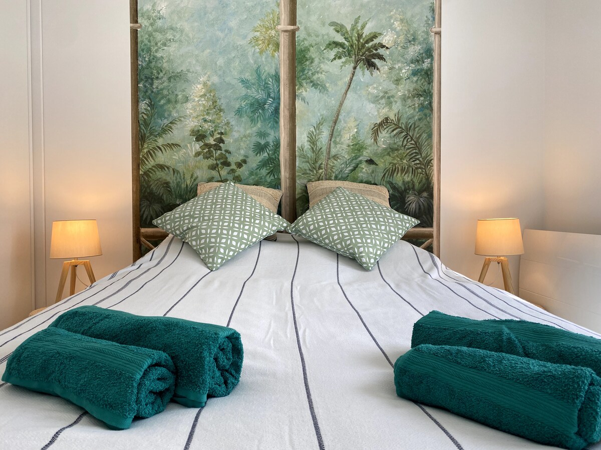 A spacious bed is centered against a wall featuring a tropical mural of lush greenery. Two patterned cushions are placed on the bed, and folded teal towels are arranged neatly at the foot. Soft lighting from two bedside lamps enhances the calming atmosphere.