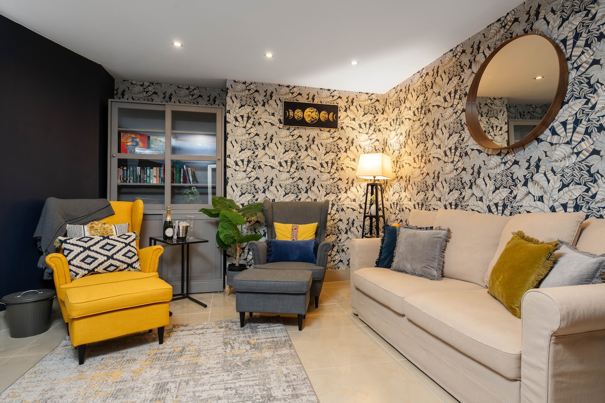 A cozy lounge area features a light-colored sofa and a comfortable armchair with yellow upholstery. A patterned rug and potted plant enhance the space, while a round mirror reflects the room's natural light. Soft lighting is provided by a standing lamp.