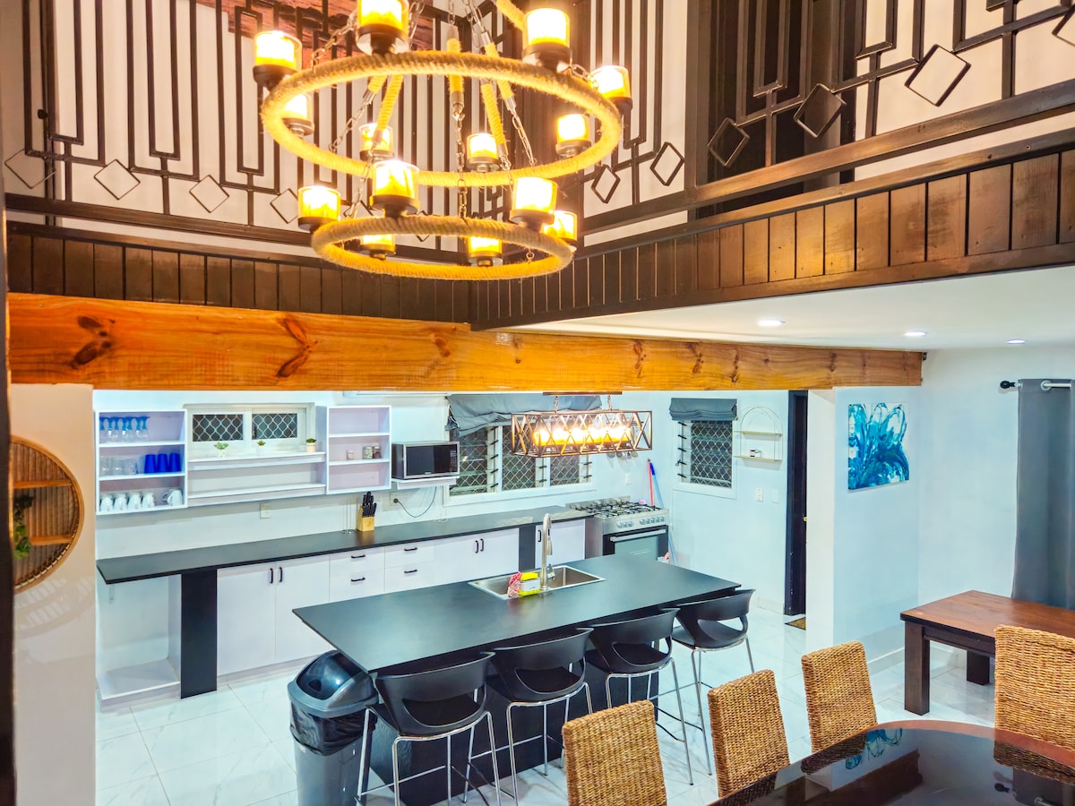 An open-plan kitchen and dining area features modern furnishings, including a large central island with a built-in sink and seating. A stylish light fixture hangs from the ceiling, illuminating the space. The kitchen is equipped with ample storage and appliances, contributing to a welcoming atmosphere.