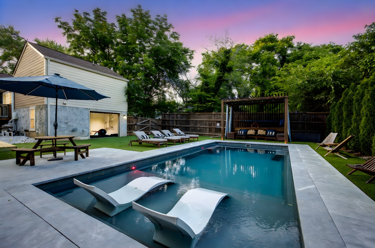 A private pool is showcased with built-in loungers and soft lighting, surrounded by a fenced yard with astroturf. Sun loungers are positioned along the poolside, and comfortable seating is available under a shaded area. Lush greenery is visible in the background.