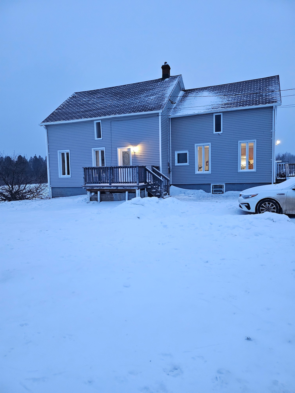 A two-story house is portrayed in a snowy setting, featuring a light grey exterior and a welcoming front porch. Large windows illuminate the home, contrasting with the wintry landscape. A vehicle is parked nearby, and the surrounding area is blanketed in fresh snow.