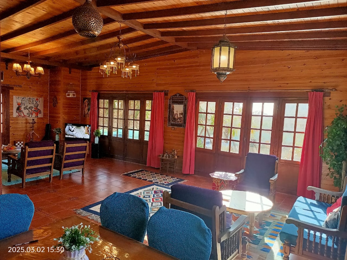 A spacious living area features wooden beams along the ceiling and warm-toned walls. Large windows allow natural light to filter in, highlighting the cozy seating arrangements. Colorful curtains frame the windows, and decorative rugs add texture to the floor.