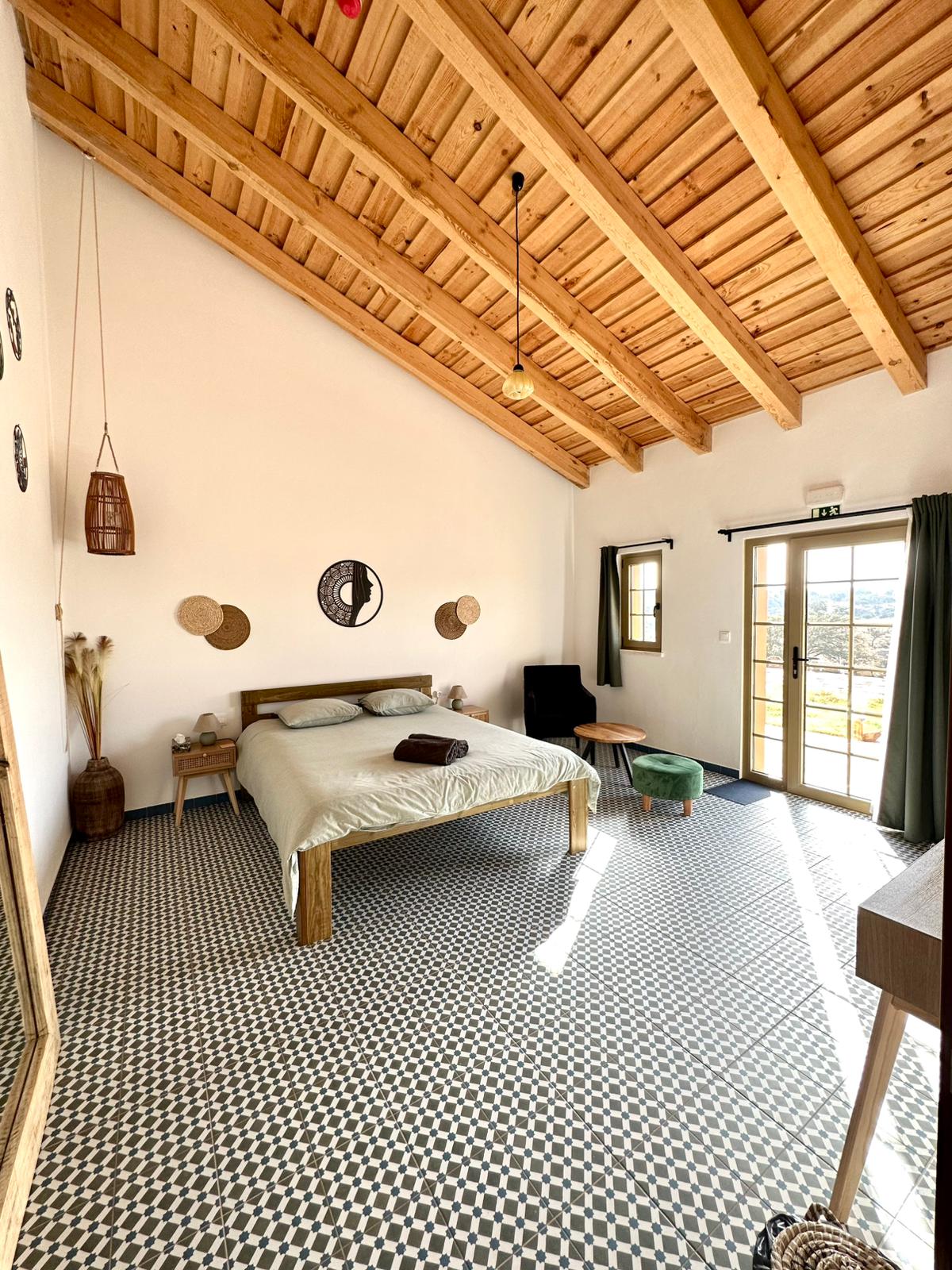 The room features a spacious bed with a neutral-colored blanket, positioned under a vaulted wooden ceiling. Natural light enters through the glass door, complemented by patterned tile flooring. Decorative wall accents and a small seating area create a welcoming environment.