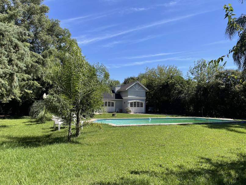 A spacious backyard is highlighted by a well-maintained lawn and a large swimming pool. The house is visible in the background, surrounded by trees that provide shade. Clear blue skies enhance the outdoor setting, creating a serene environment.