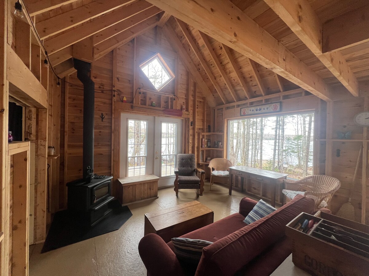 The inviting living space features a wood stove, wooden beams, and large windows offering scenic views of the lake. A comfortable sofa and armchairs are positioned around a coffee table, enhancing the cozy atmosphere. Natural light floods in through the diamond-shaped window.