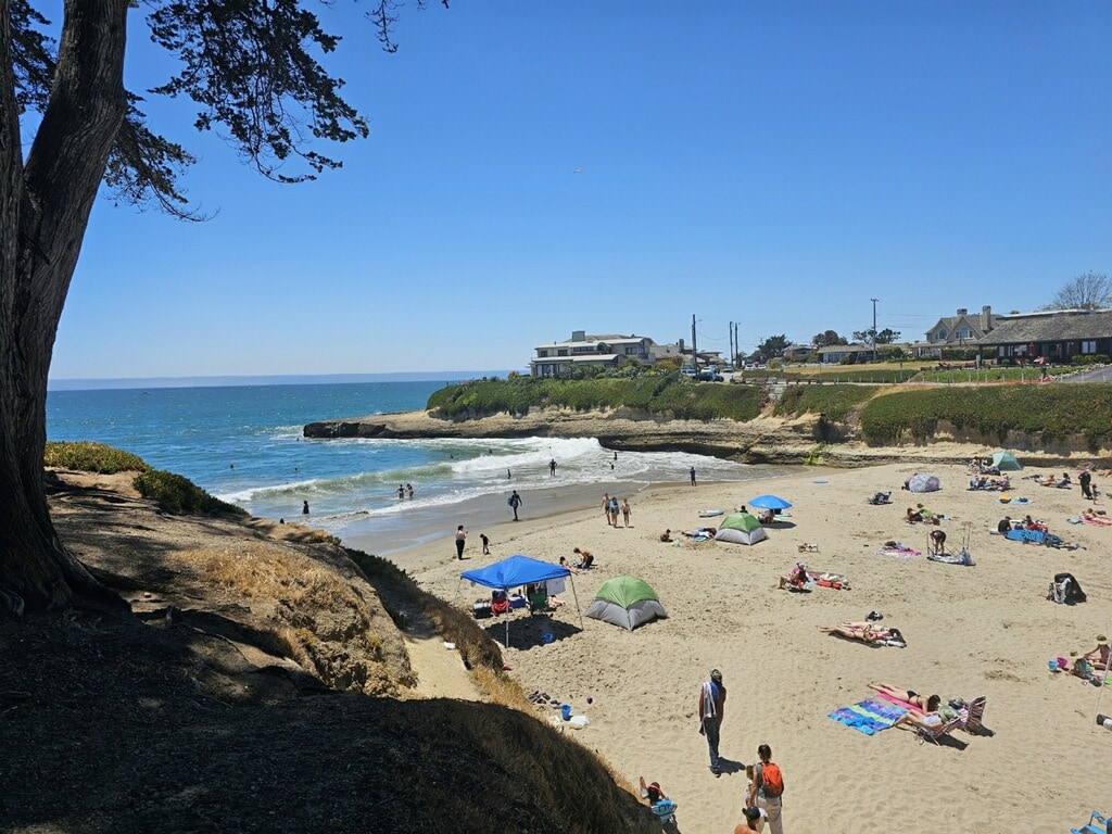 A sandy beach scene captures visitors enjoying the sun, with colorful umbrellas dotting the shore. Gentle waves lap at the coastline, and the blue ocean stretches into the distance. Cliffs and greenery frame the beach, creating a serene coastal environment.