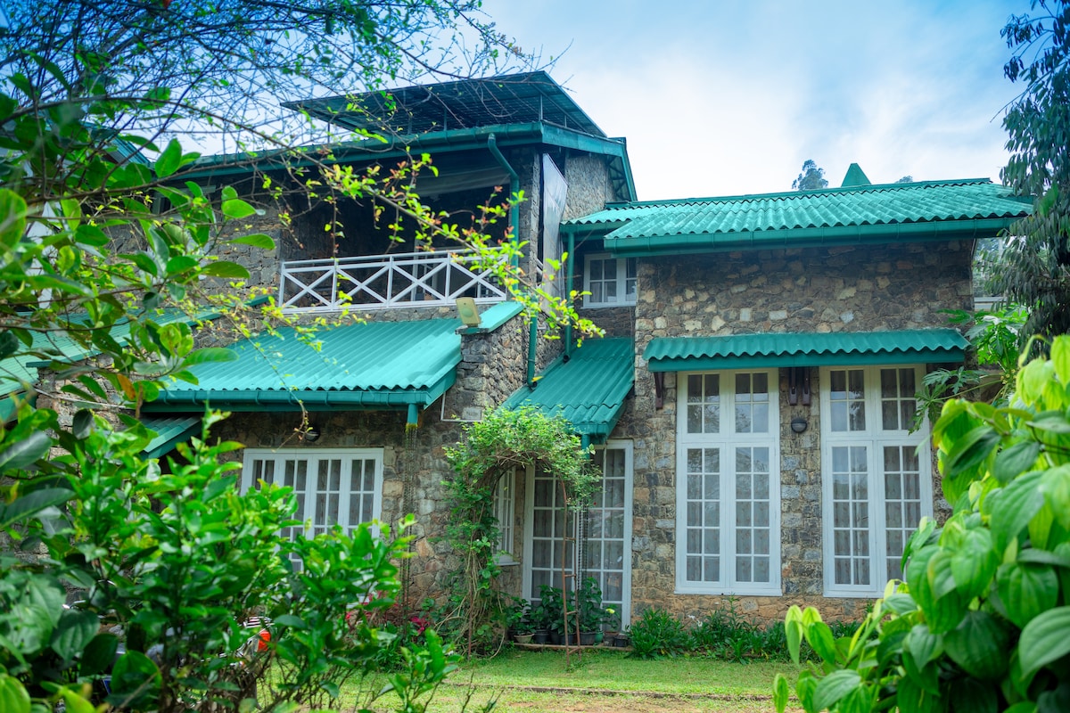 The villa features a charming stone facade with green roofing, surrounded by lush foliage. Large windows allow natural light into the structure, which is set within a vibrant garden. Architectural details reflect a colonial tea planter's style while blending harmoniously with the scenic surroundings.