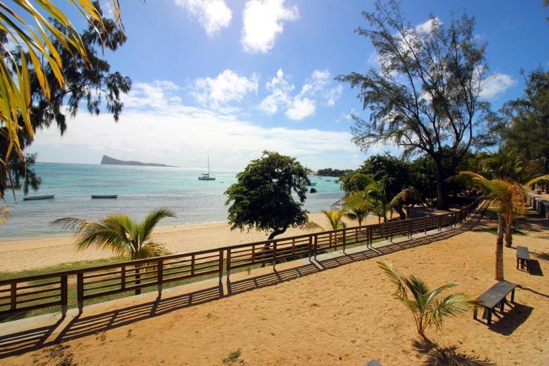 A sandy beach extends under a clear blue sky, framed by palm trees. Gentle waves lap at the shore, while a few boats are visible on the water. A wooden railing lines the pathway, providing a natural border between the beach and the lush greenery.