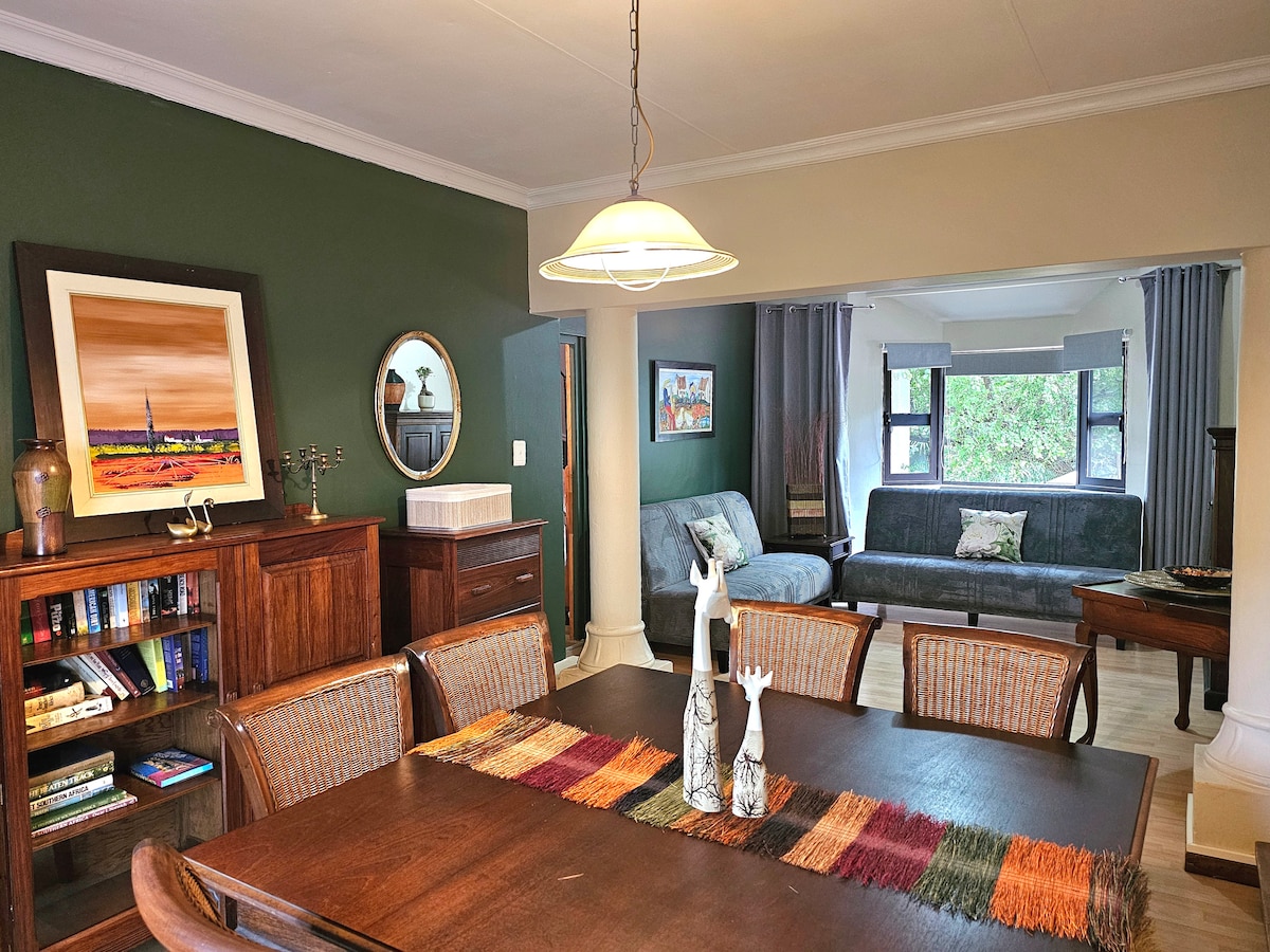 A shared living space is featured with a large wooden dining table adorned with a colorful runner. Light green walls are complemented by wooden furniture. A comfortable lounge area contains a fold-out couch and large windows, allowing natural light to fill the room.