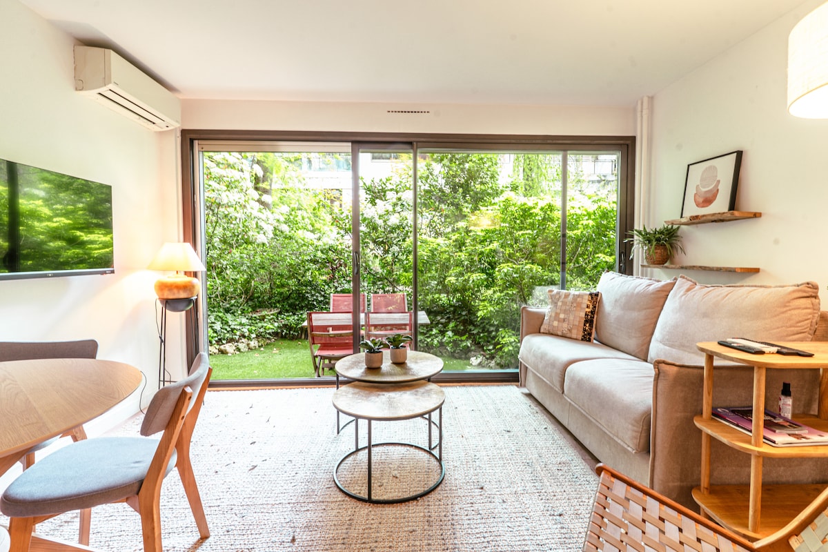 A living area features a comfortable sofa and a round coffee table set on a textured rug. Large glass doors open to a lush garden, enhancing natural light. A dining table with chairs is positioned nearby, while decorative plants and warm lighting add a cozy touch.