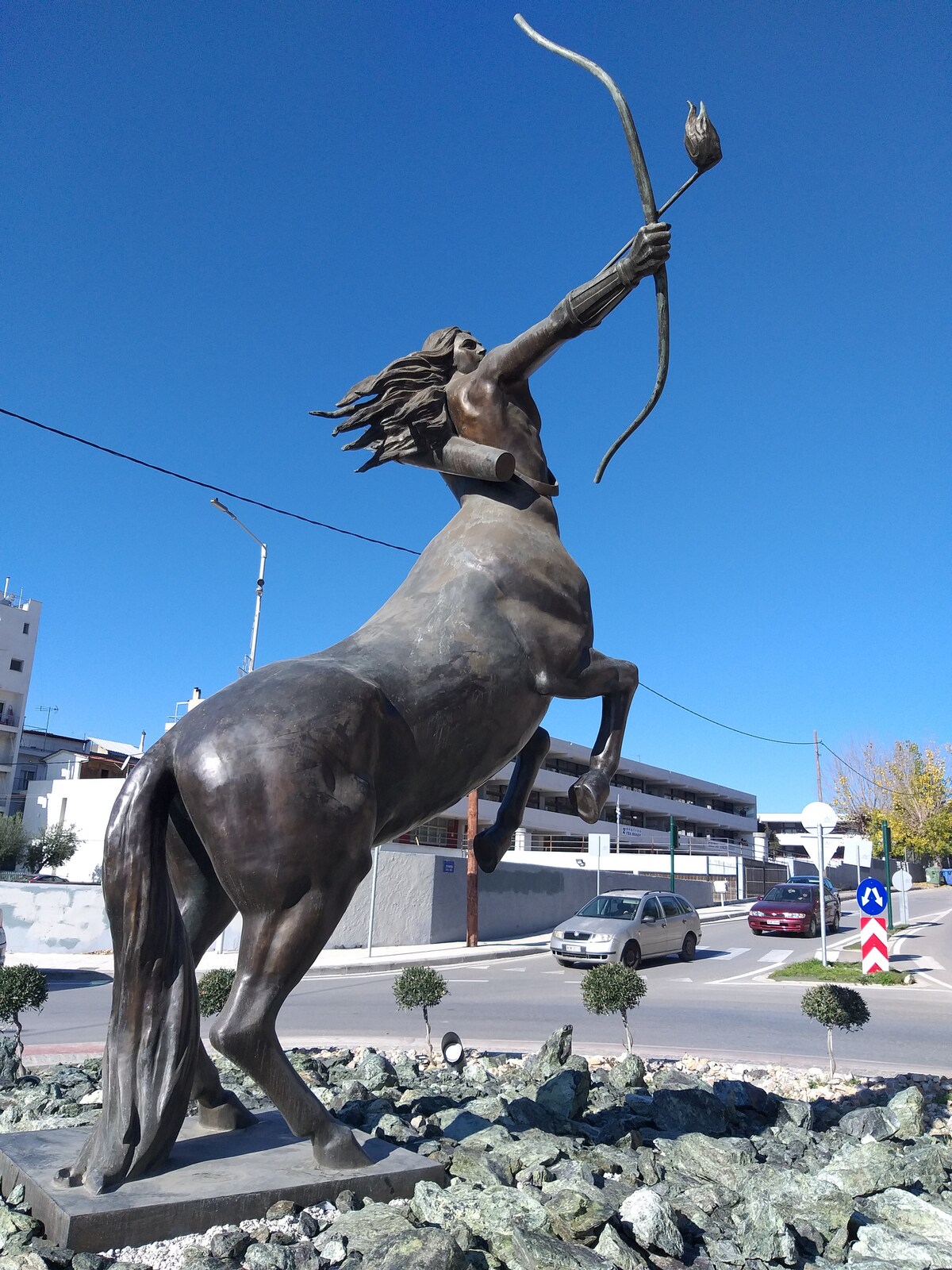 A striking bronze statue of a centaur stands tall, capturing a dynamic pose with a raised bow. The figure is set against a clear blue sky, surrounded by smooth stones and small shrubs, conveying a sense of strength and artistry at a public intersection.