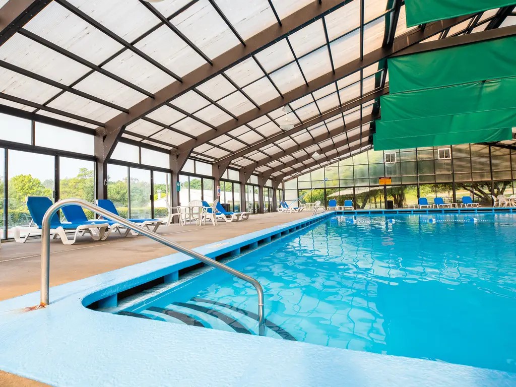 An indoor pool area features a clear blue water surface, surrounded by ample seating with lounge chairs. High ceilings and large glass panels allow natural light to illuminate the space, with views of the outdoors visible through the windows.