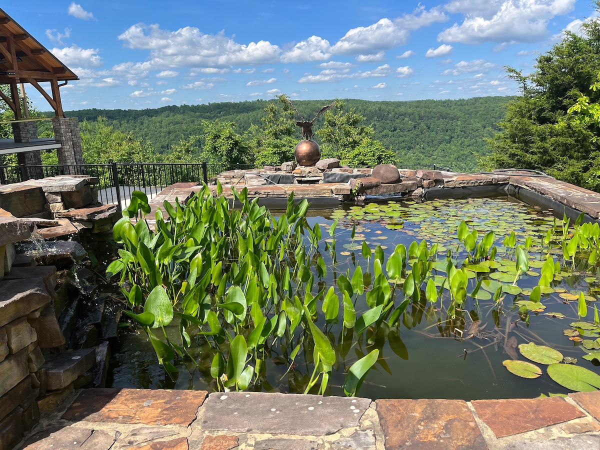 A tranquil Koi pond is surrounded by natural stone, featuring lily pads floating on the surface. The clear sky is visible above, with lush green mountains in the background, completing the serene outdoor setting.