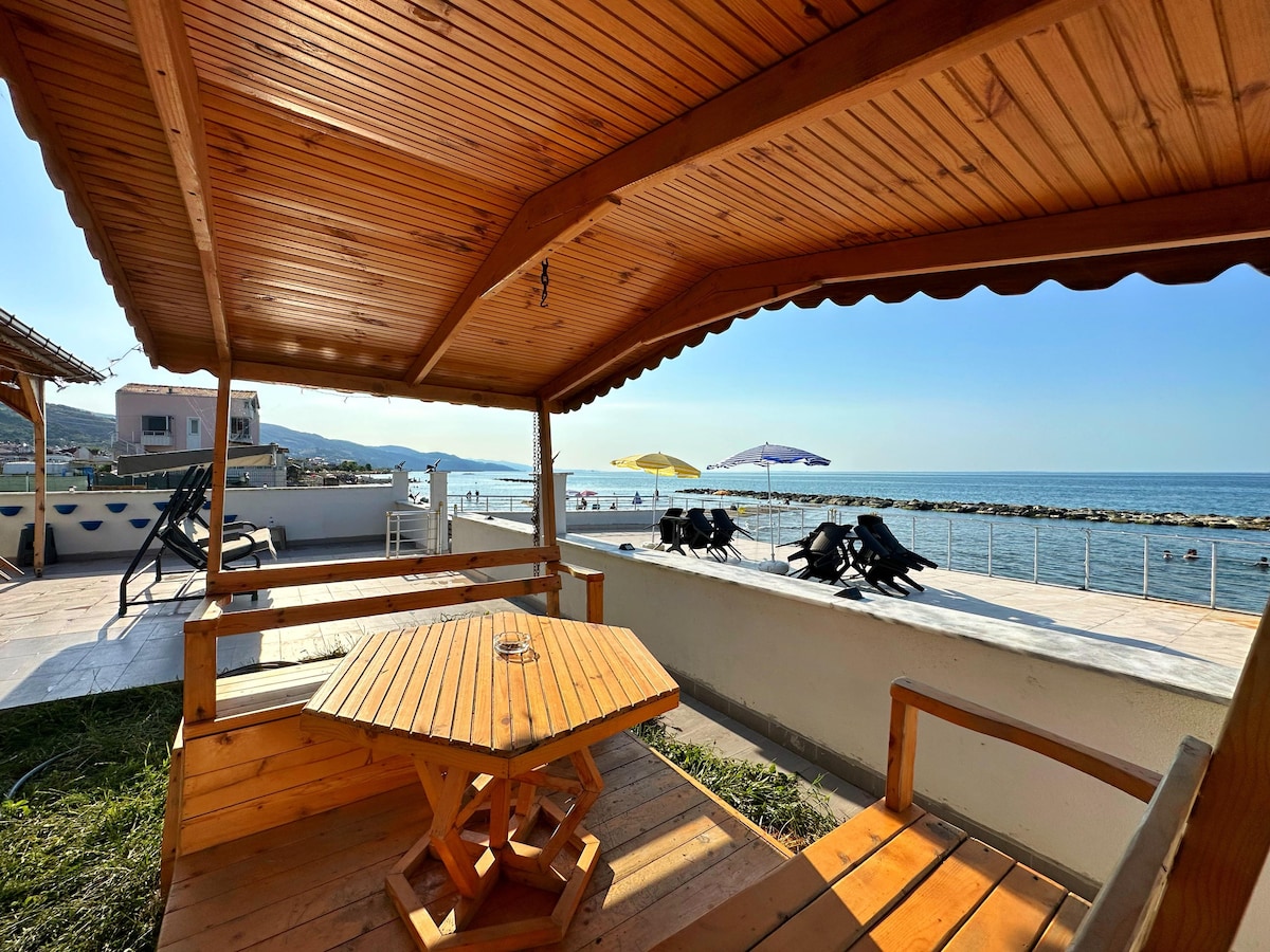 A wooden gazebo with a hexagonal table provides shaded seating overlooking a pebbled beach. Sun loungers and umbrellas are visible along the shoreline. The calming sea is just beyond, with distant hills framing the tranquil view under a bright blue sky.