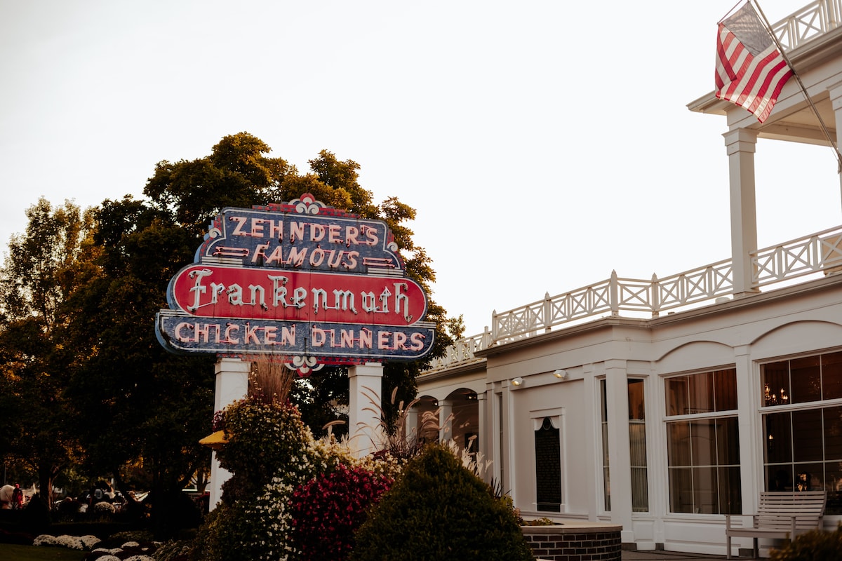 A vintage sign for Zehnder's Famous Frankenmuth Chicken Dinners is prominently displayed in front of a well-maintained building. The architecture features large windows and elegant outdoor decor. Lush greenery and colorful flowers surround the sign, providing a lively backdrop.
