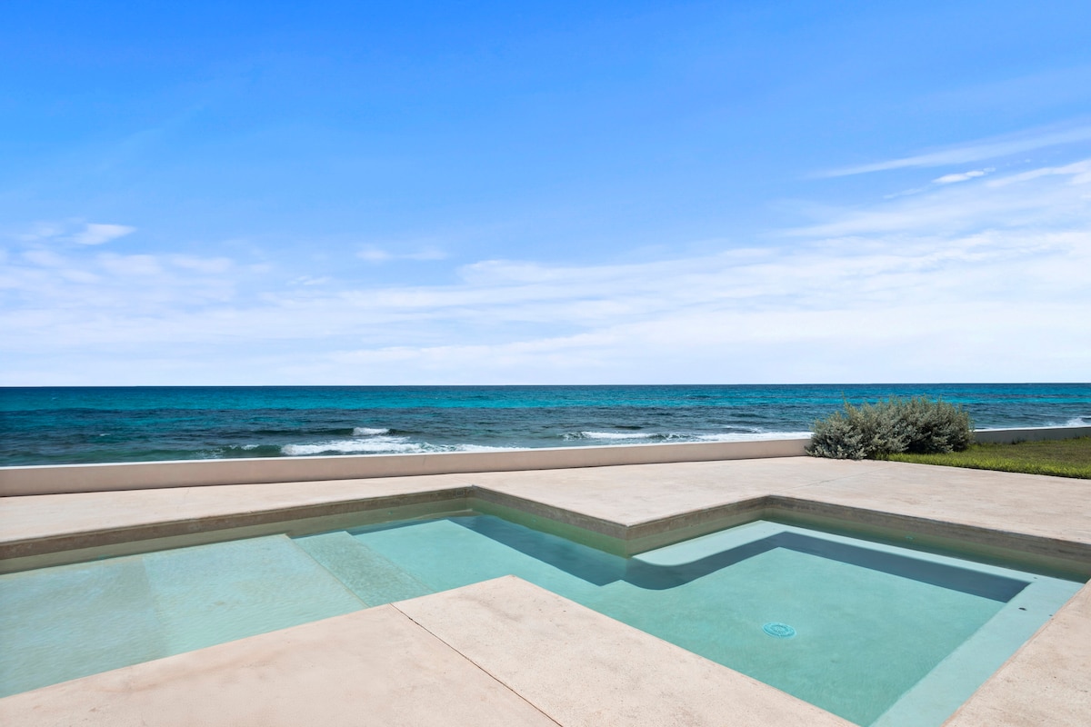 A serene view of the ocean is presented from the edge of a modern pool. The calm turquoise waters of the sea meet the horizon under a clear blue sky, while a section of well-maintained grass borders the pool area.