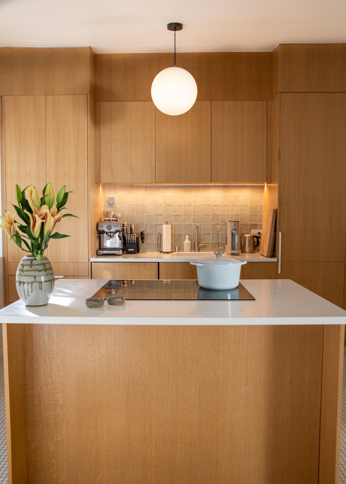 The kitchen is characterized by warm wood cabinetry and a modern design. A sleek white countertop features a blue cooking pot, while an espresso machine and an assortment of kitchen tools are visible. Fresh flowers in a vase bring a touch of nature indoors.