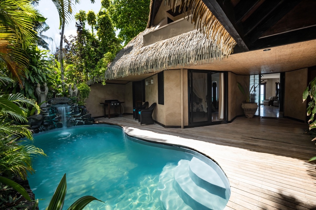 A private swimming pool with a cascading waterfall is surrounded by lush tropical gardens and wooden decking. The villa interior is visible through expansive glass doors, allowing natural light to fill the space. The thatched roof adds a touch of island charm.