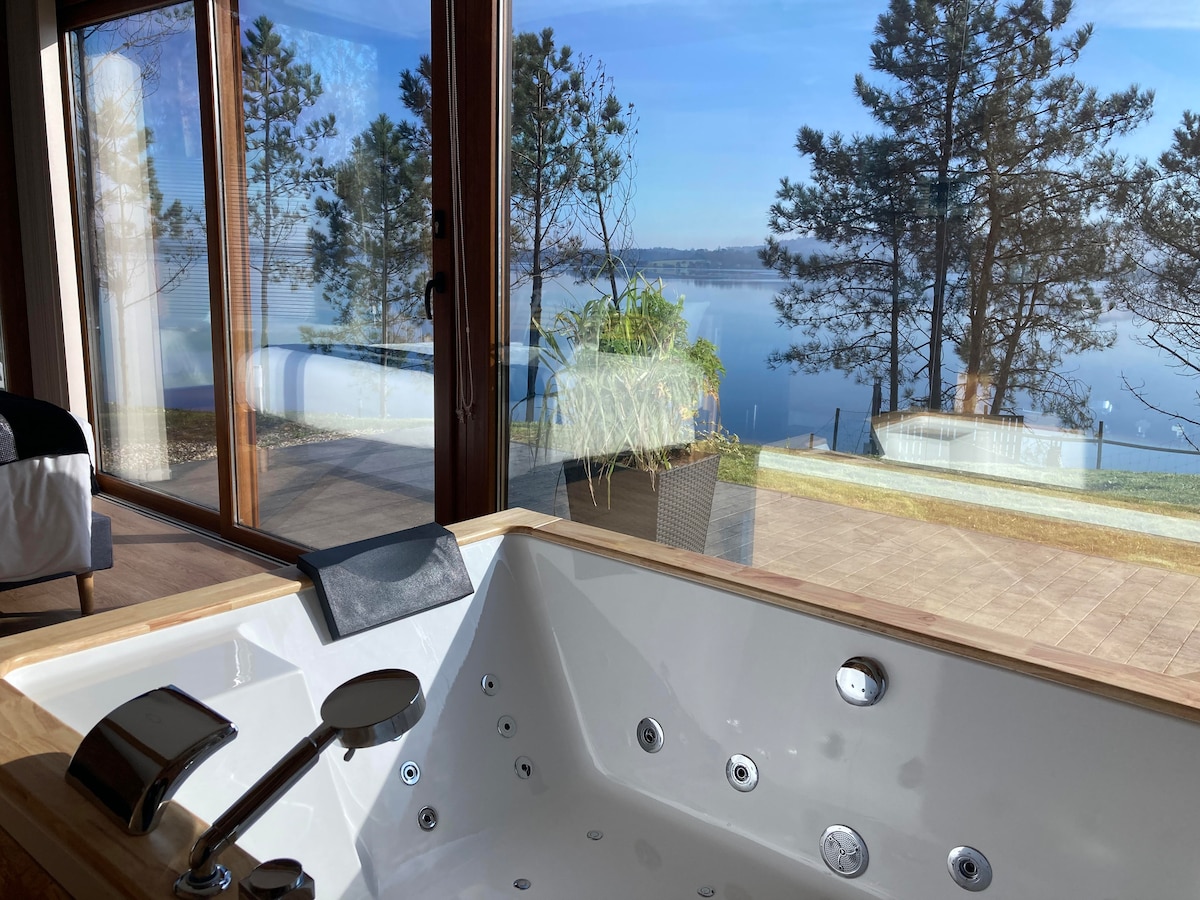 An outdoor Jacuzzi is shown in the foreground, featuring multiple jets. Large glass doors provide a view of the serene A Fervenza reservoir, with trees in the distance and calm water reflecting the sky.