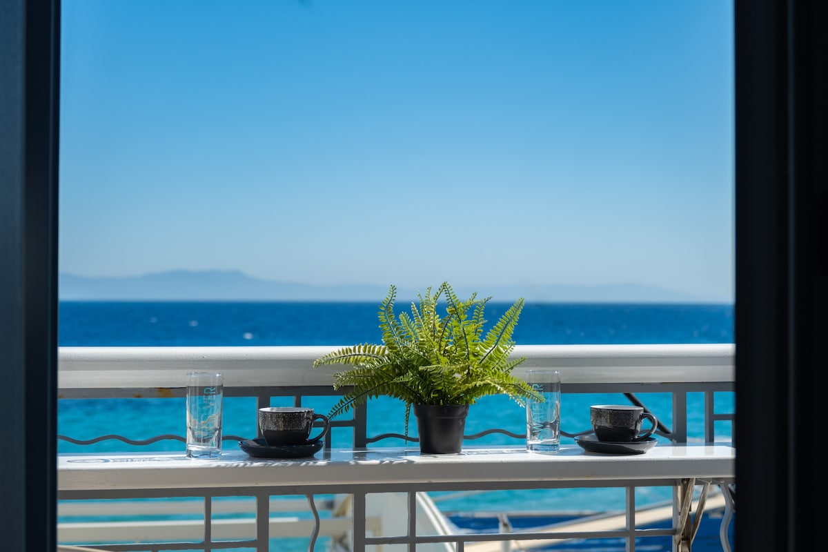 A balcony space overlooks the sea, featuring a small potted fern and two coffee cups beside clear glasses. The calm blue waters and bright sky create a serene backdrop, enhancing the inviting outdoor setting.