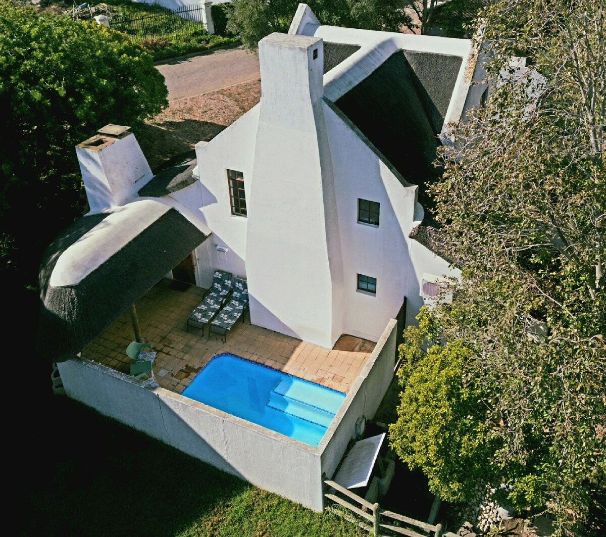 The thatched-roof home is surrounded by lush greenery and features a private pool with a tiled patio. Sun loungers are arranged by the pool, providing a relaxing space. A shaded area can be seen adjacent to the pool for outdoor dining or lounging.