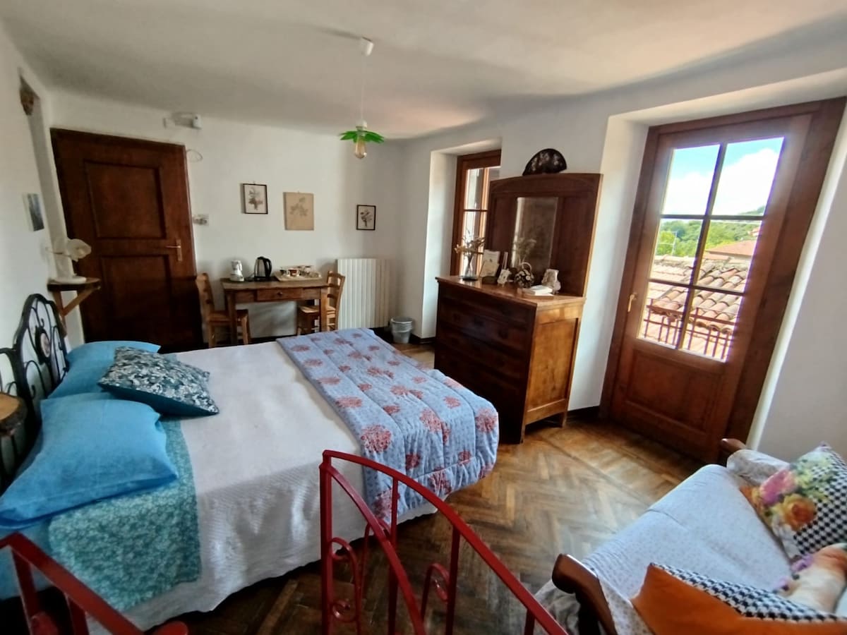 A bright room features a spacious bed layered with a colorful quilt, complemented by decorative pillows. A wooden dresser and a small seating area provide additional comfort. Large windows frame views of the surrounding hills, allowing natural light to fill the space.