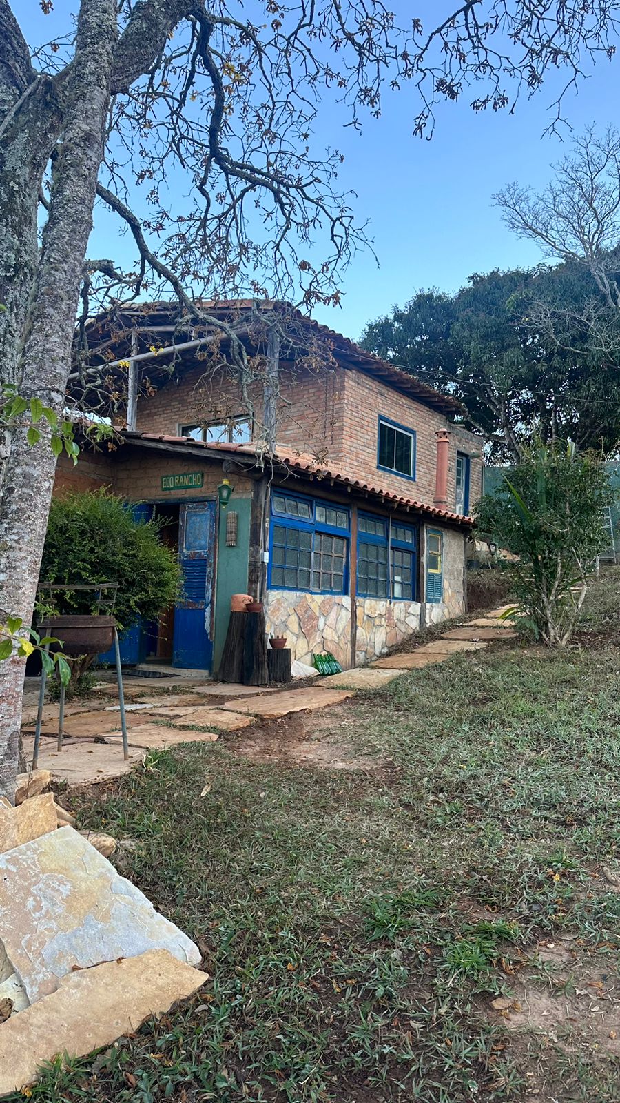 A rustic house is set against a natural backdrop, constructed with repurposed materials. The two-story structure features blue-framed windows and a terracotta roof. Lush greenery lines the pathway leading to the entrance, with decorative elements visible by the door.