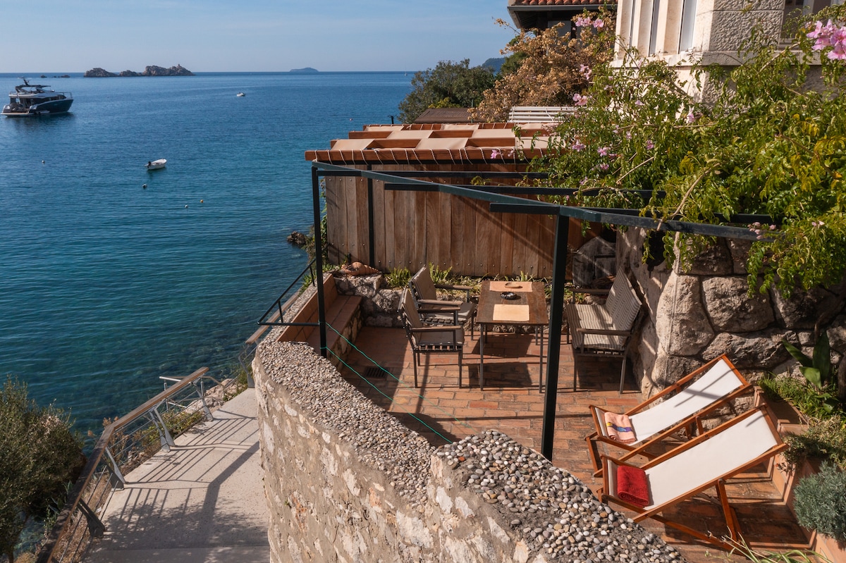 A private terrace is presented with a seating set and lounge chairs, overlooking the tranquil sea. Lush greenery adds a natural touch, while the sunlit area invites relaxation. Steps lead down towards the rocky beach, enhancing the close proximity to the water.