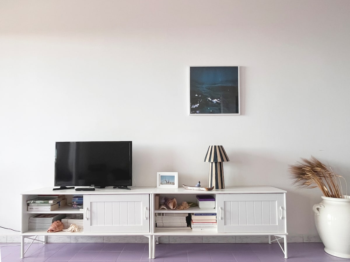 A modern living area is presented, featuring a white media console with a television and decorative items. A framed artwork is displayed on the wall above. A striped lamp and a decorative vase with dried plants add contrasting textures to the space.