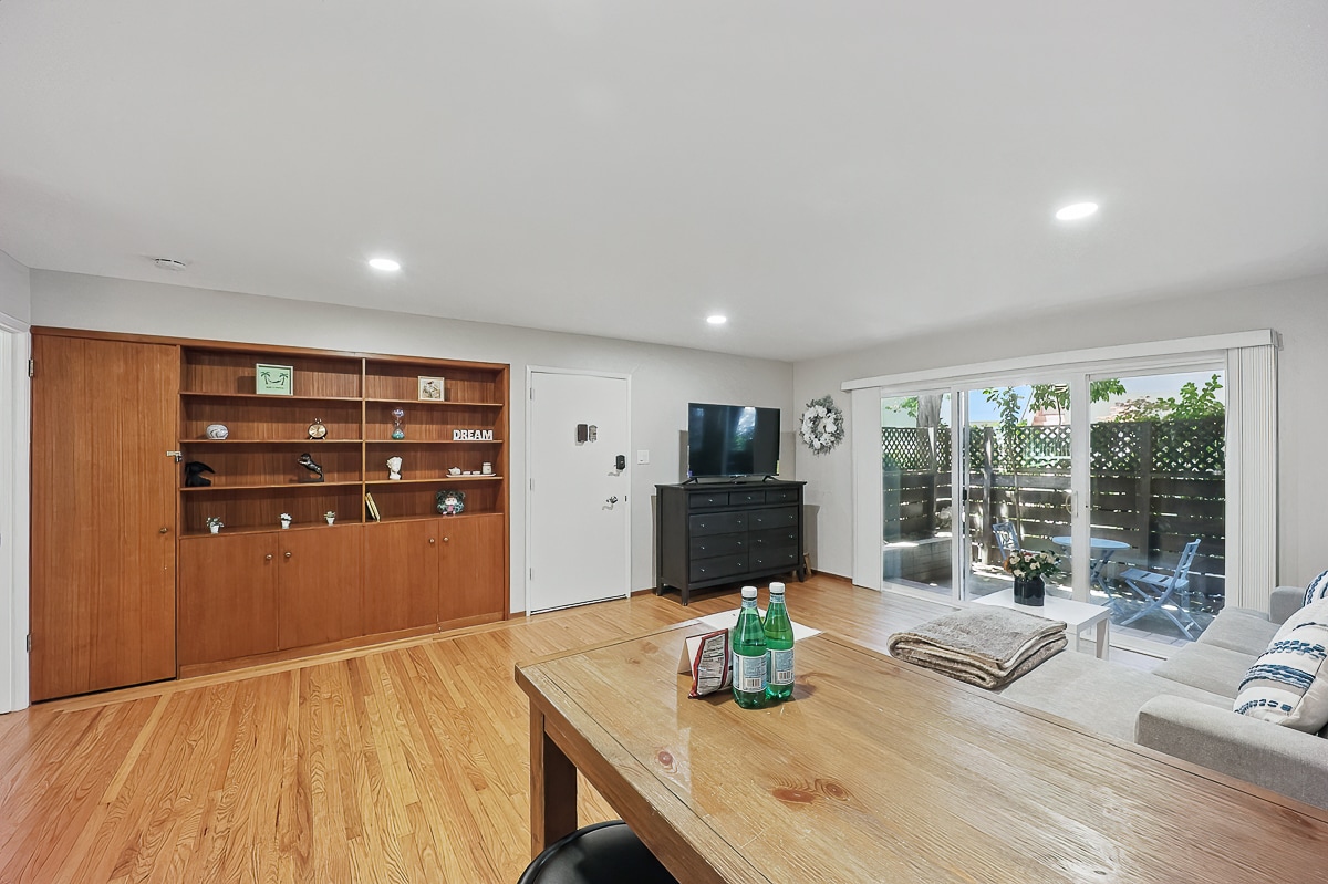 A spacious living area features warm hardwood flooring and built-in wooden shelving displaying decorative items. Large sliding glass doors lead to a private fenced patio, allowing natural light to fill the room. A dark wood dresser and a smart TV complete the comfortable ambiance.