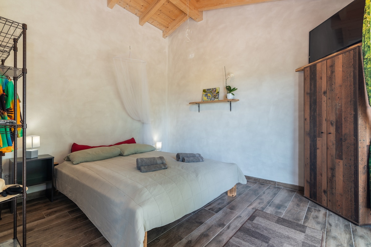 A spacious sleeping area features a large bed with soft bedding, flanked by contemporary lamps on either side. A rustic wooden shelving unit holds decorative items, and a TV is mounted on the wall. Light enters through a window draped with a sheer curtain.