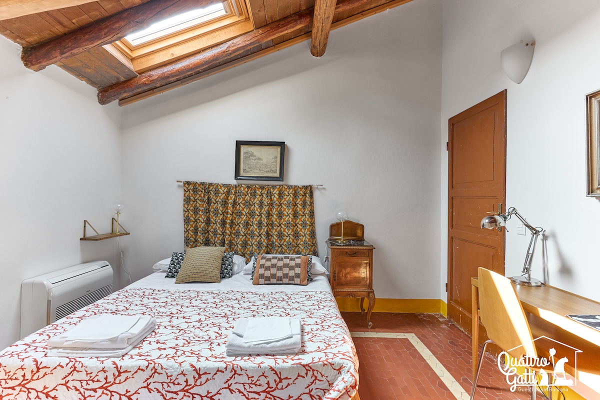 A comfortable room is presented with a spacious bed adorned with decorative pillows. Wooden ceiling beams and a skylight enhance brightness. A vintage bedside table and a writing desk complete the setting, providing functional spaces. Fresh linens are neatly placed at the foot of the bed.