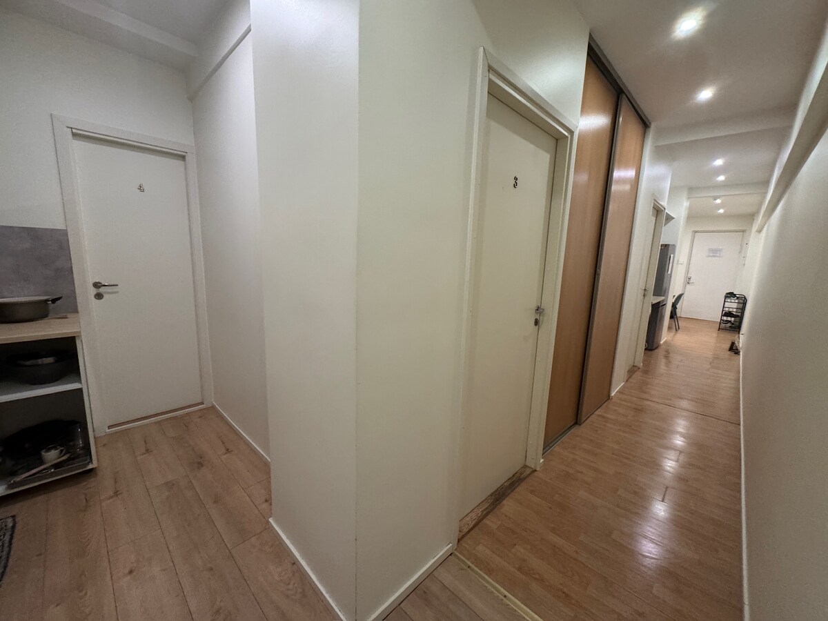 A well-lit hallway features smooth wood flooring and white walls. Multiple doors are visible, indicating private rooms. A small storage area with visible shelves and a kitchen space is situated on the left, while more doors line the corridor on the right.