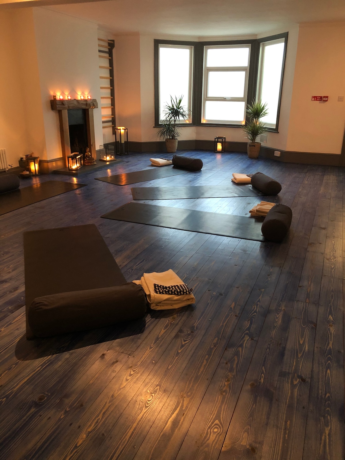 A serene wellness space features multiple yoga mats arranged on a rich wooden floor. Soft lighting from candles and lanterns creates a calm atmosphere, while potted plants add a touch of nature. Floor cushions and folded towels are positioned on the mats for comfort.