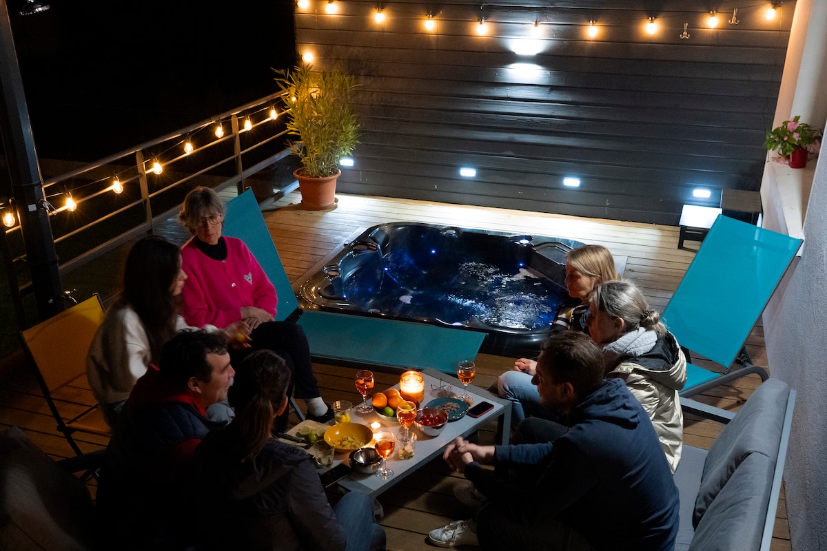 A relaxing outdoor space is showcased, featuring a hot tub surrounded by soft lighting. Comfortable seating is arranged around a central table, which holds plates of food and drinks. The atmosphere is intimate and designed for gatherings, with plants adding natural elements.