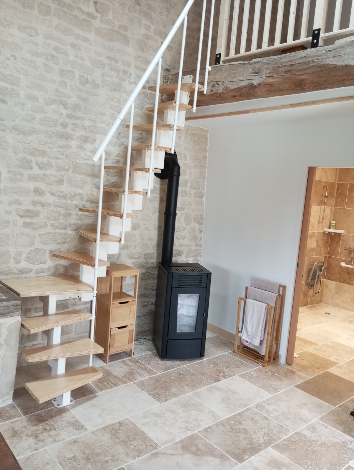 A spiral staircase with wooden steps leads to the mezzanine area. A modern pellet stove is positioned against a stone wall, providing warmth. The tile flooring features a mix of light and darker tones, complementing the neutral color palette of the space.
