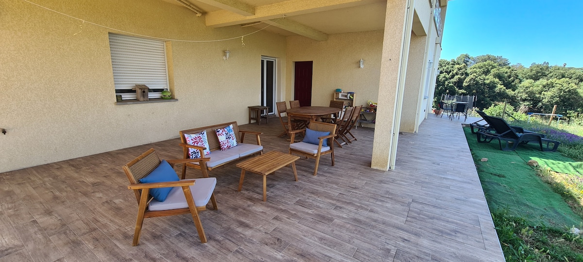 A spacious terrace is showcased with multiple seating options, including wooden chairs and a central table. A dining set is present in the background, surrounded by greenery. Natural light fills the area, and a scenic view is visible beyond the patio.