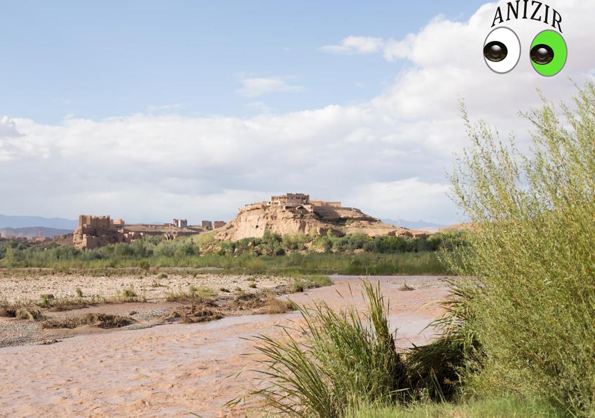 A scenic view showcases a historic structure atop a rocky outcrop, surrounded by greenery. A gently flowing river meanders in the foreground, with tall grass adding texture to the landscape. The sky holds a mix of clouds and sunlight, creating a peaceful atmosphere.