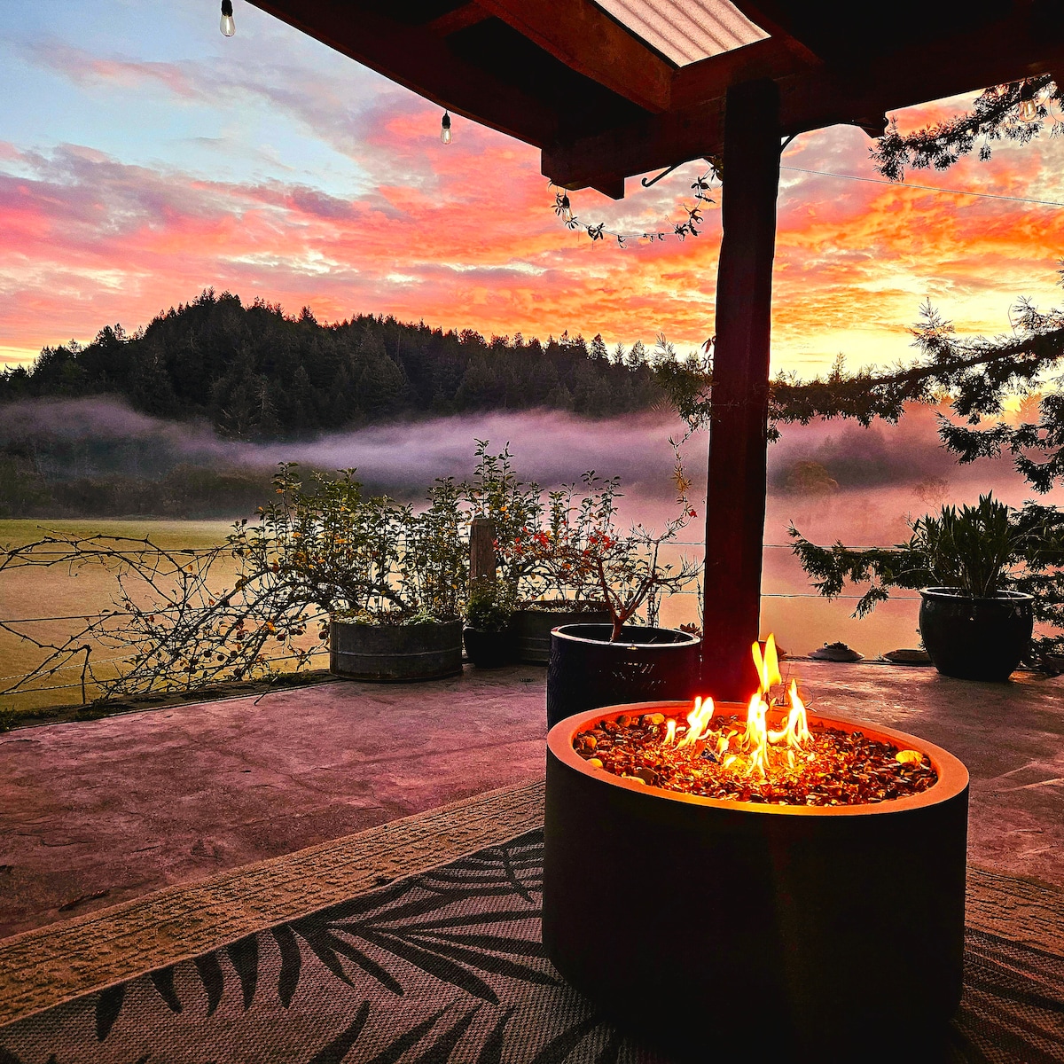 A fire pit is centrally located on a cozy back patio, with flames casting a warm glow. Lush greenery surrounds the area, while a foggy meadow and forest backdrop are illuminated by a vibrant sunset sky in shades of orange and pink.