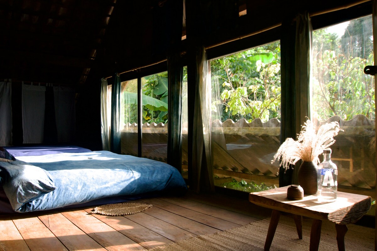 A spacious sleeping area features a large bed with soft indigo bedding. Two expansive glass doors invite views of lush greenery outside. Natural light filters through sheer curtains, illuminating the rustic wooden floor and a small table adorned with decorative items.