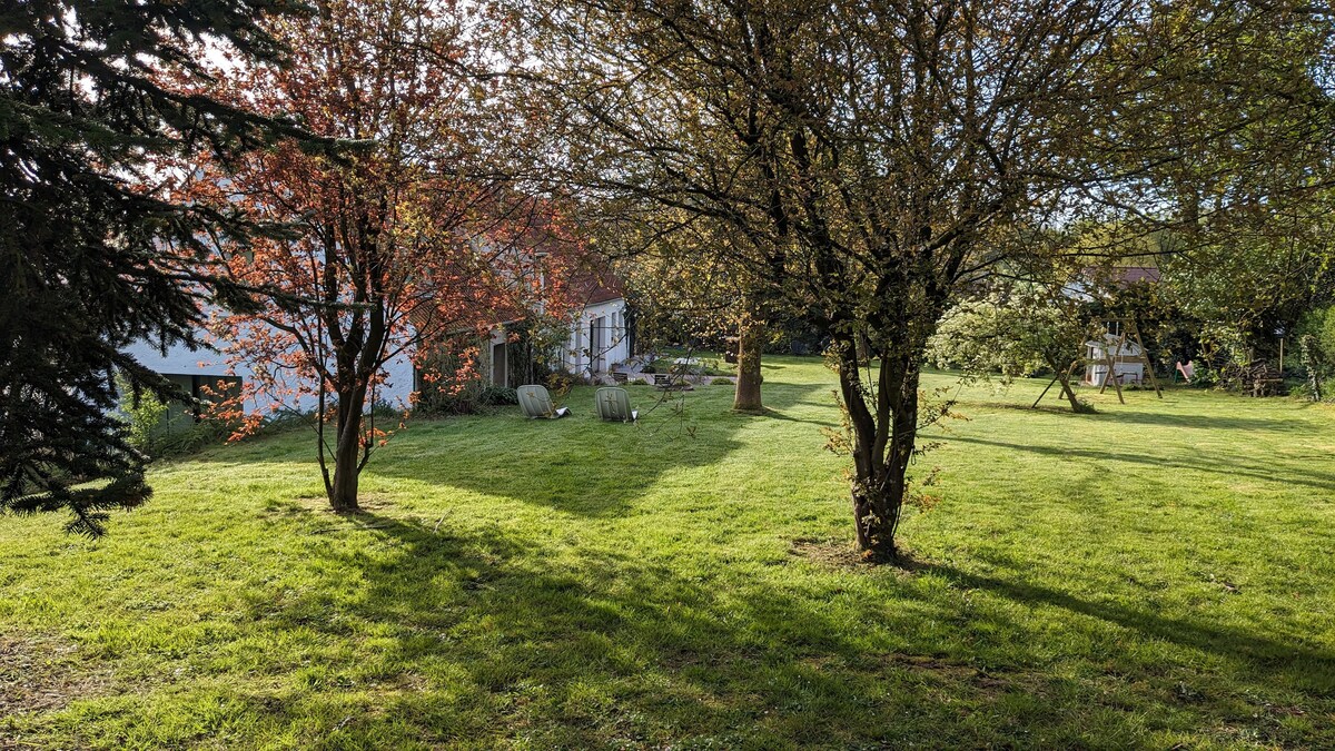 The spacious garden features well-maintained grass and several trees that provide shade. Two stone benches are positioned under the trees, creating a quiet outdoor seating area. The landscape is open, with a glimpse of the house set against a serene backdrop.