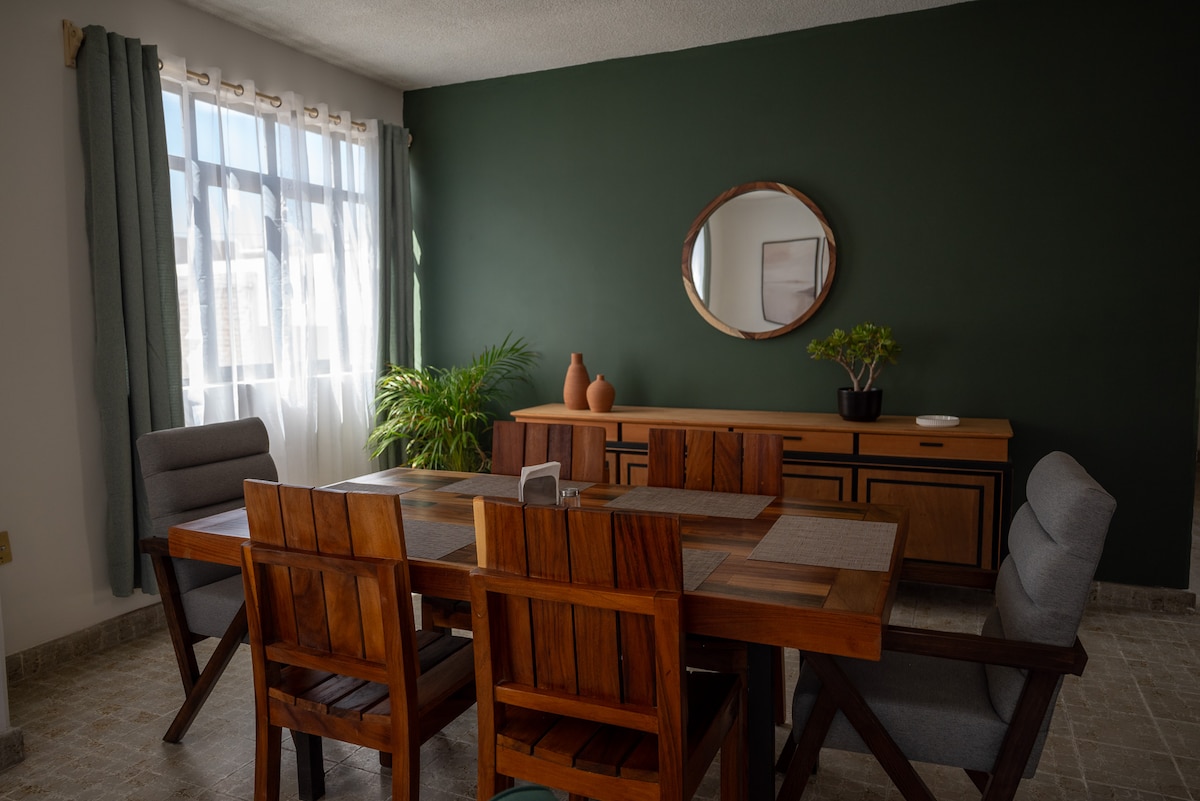 A dining area features a large wooden table surrounded by six chairs. A mirror is mounted on a green wall above a low sideboard. Natural light streams through large windows, illuminating a potted plant positioned in the corner.