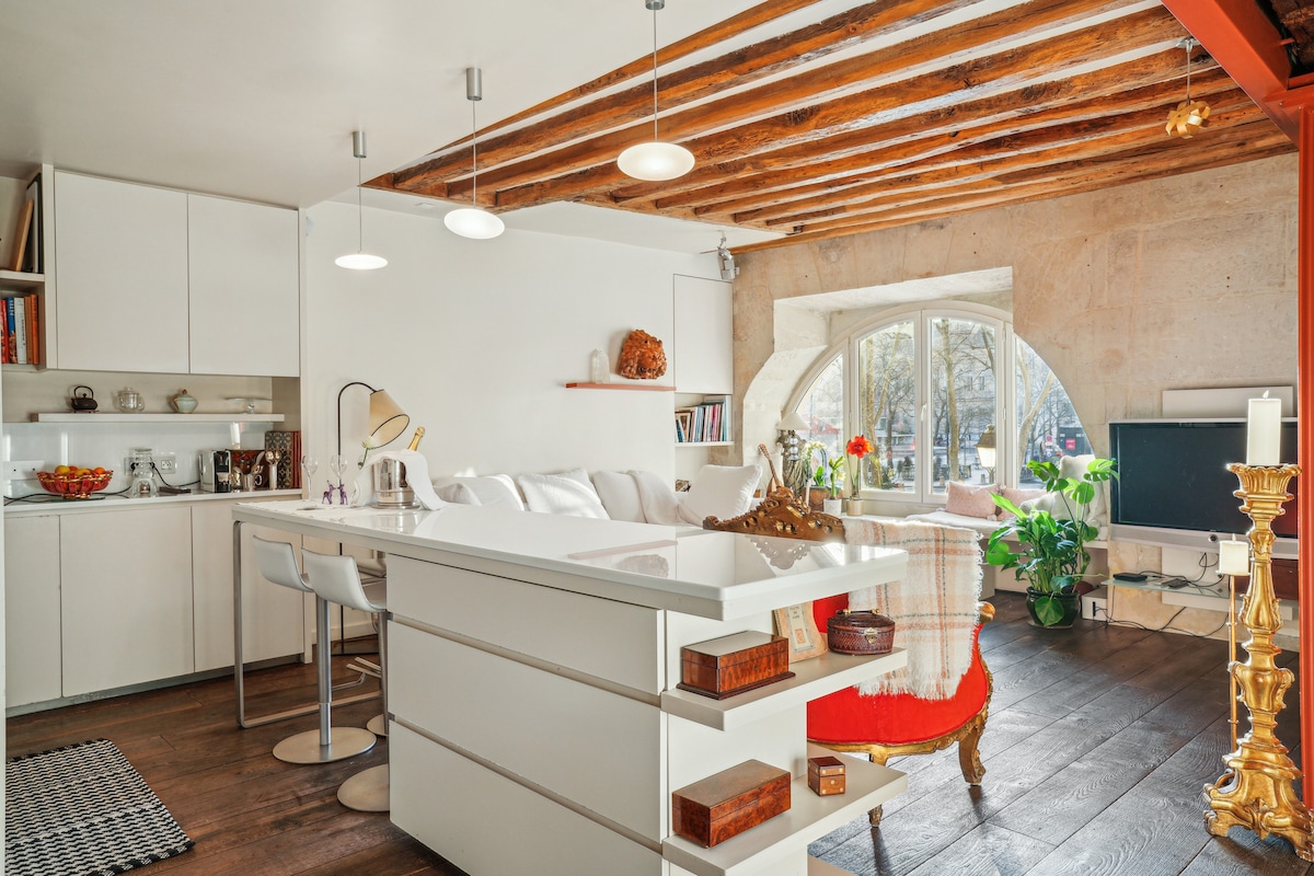 A spacious living area features a modern kitchen with an open layout, showcasing white cabinetry and a central island. Natural light floods through large curved windows, illuminating comfortable seating and greenery. Exposed wooden beams add character to the space.